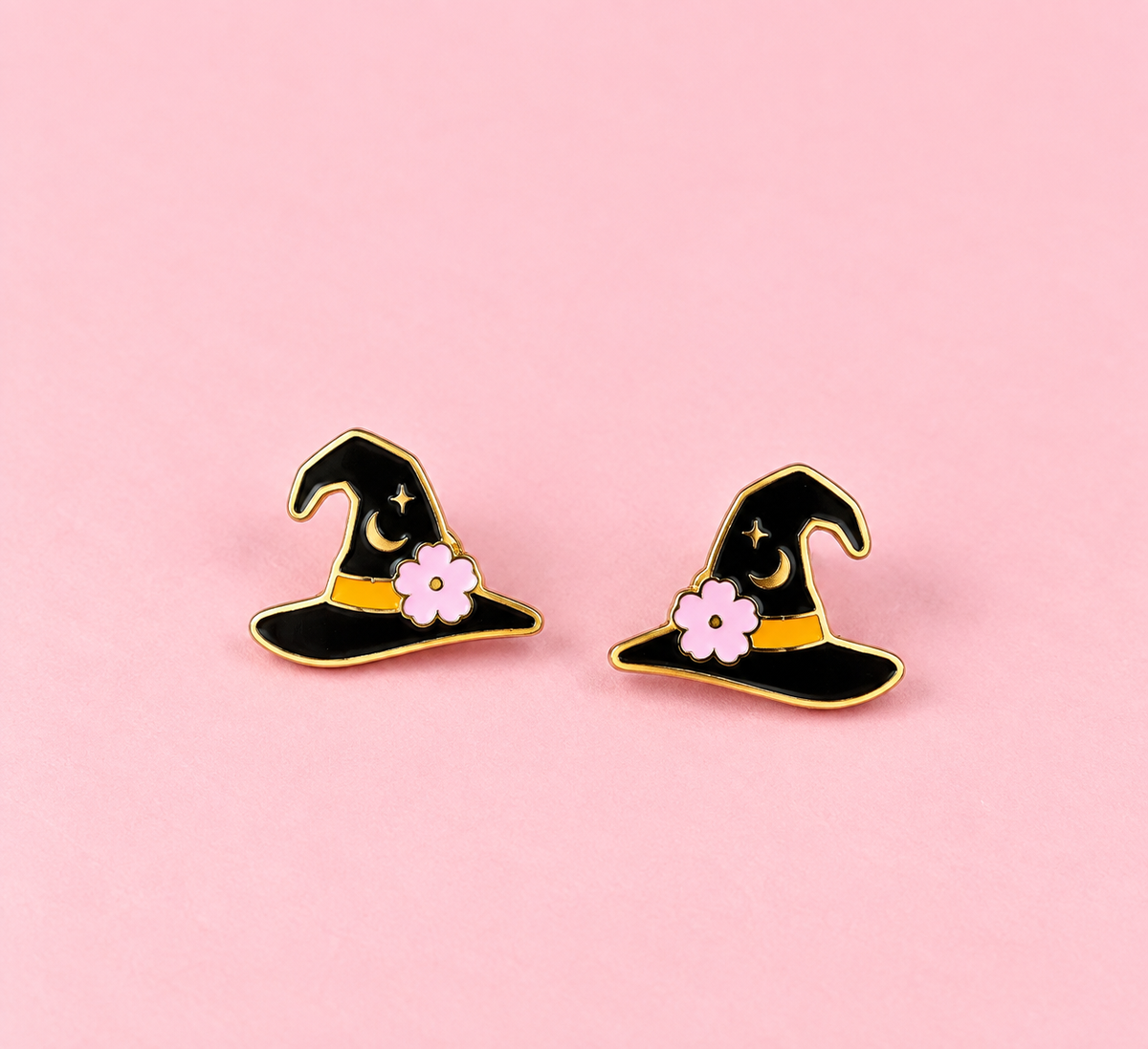 A pair of witch hat stud earrings with black enamel, gold metal trim, a small pink flower, and a gold crescent moon, displayed on a solid pink background.