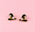 A pair of witch hat stud earrings with black enamel, gold metal trim, a small pink flower, and a gold crescent moon, displayed on a solid pink background.