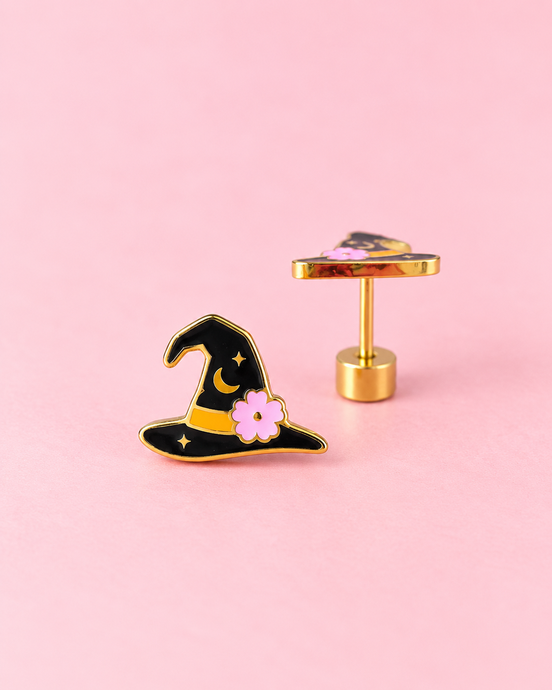 A pair of gold-tone earrings shaped like a witch's hat, featuring black enamel, a yellow band, and a pink flower, displayed on a solid pink background.