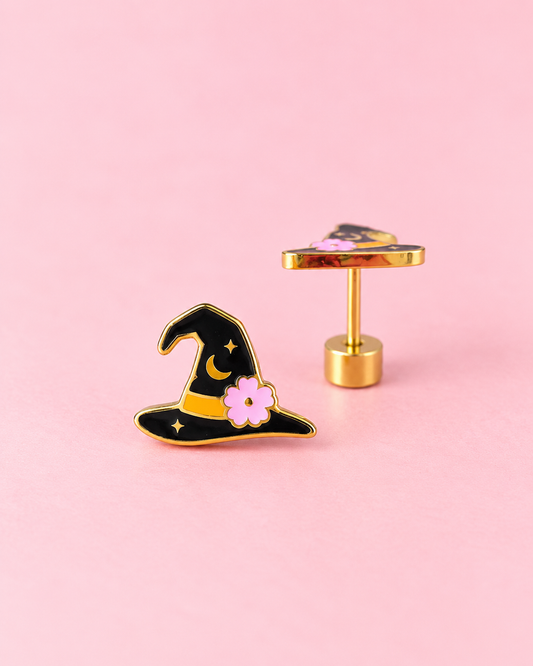 A pair of gold-tone earrings shaped like a witch's hat, featuring black enamel, a yellow band, and a pink flower, displayed on a solid pink background.